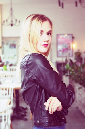 Beautiful young blonde woman posing in cozy cafe. She wears a black leather jacket, black lacy tops and blue jeans. Large bright windows in the cafe. Sexual posture.の写真素材