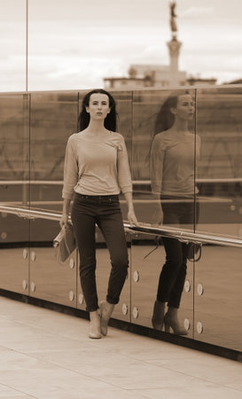 Pretty young woman posing near a glass wall in the downtown. She is wearing burgundy jeans, pink shirt and yellow shoes, with yellow handbag in her right hand.の写真素材