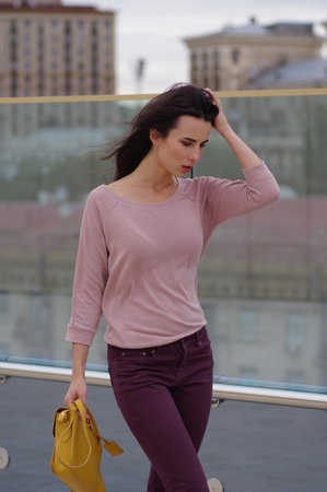 A young woman poses near a glass wall in the city. Street dress style. She wears Burgundy jeans, a pink shirt. Looking downの写真素材