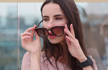 A long-haired young brunette woman posing while playing with sunglasses. Fashionable style with pink tones. Blurred background. The wind plays with brunette hair.の写真素材