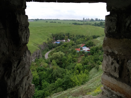 A pastoral rural landscape with green hills and a meadow, a cleft, a mouth of a dried river, where small houses are located, sky, summer, tourist attraction,framed by old castle bricks windowの写真素材