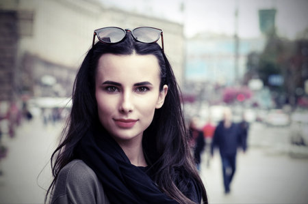 A black-haired girl in the city on the background of the avenue, passers-by, cars, in a big black scarf, glasses on her head, a jacket, street fashionの写真素材