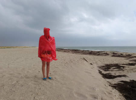 Woman in red raincoat at sea beach at windy rainy weather standing on sand, no face visibleの写真素材