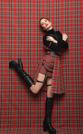 Modern fashion. A readhead girl in a plaid (checkered) dress on a plaid background in black golf with a bare leg In high boots demonstrates a modern stylish fashionable pose.の写真素材