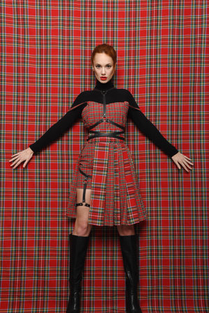 Modern fashion. A readhead girl in a plaid (checkered) dress on a plaid background in black golf with a bare leg In high boots demonstrates a modern stylish fashionable pose.の写真素材