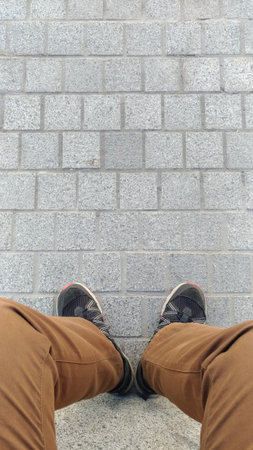 Legs of a seated man in shabby trousers and sneakers standing on a surface of paving slabs, top view, street, pedestrian zone.の写真素材