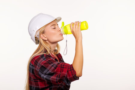 A girl in a white construction helmet and a red shirt is drinking water from a worn bottleの写真素材