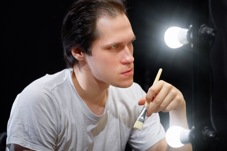 the actor sits in front of a mirror in a white t-shirt in a dark room and looks at his reflection, holding a brush for applying makeup to his faceの写真素材
