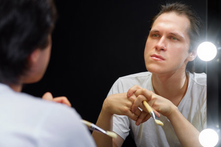 the actor sits in front of a mirror in a white t-shirt in a dark room and looks at his reflection, holding a brush for applying makeup to his faceの写真素材