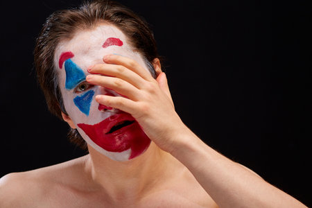 a young man smears clown makeup on his face with one hand, with bare shoulders, emotionally expresses feelings of mood, on a black backgroundの写真素材