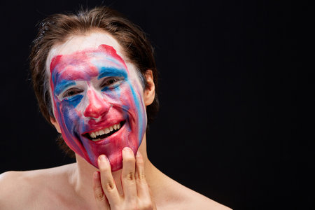 a young man smears clown makeup on his face with one hand, with bare shoulders, emotionally expresses feelings of mood, on a black backgroundの写真素材