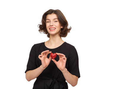 A beautiful brunette girl in a black dress on a white background holds a red decorative heart in her handsの写真素材