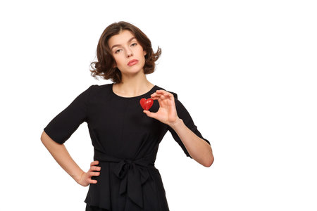 A beautiful brunette girl in a black dress on a white background holds a red decorative heart in her handsの写真素材