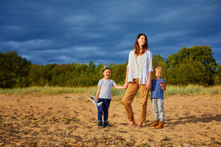 A young happy mother with two boys playing on the sand near the forest, in the hands of one of them a model of a civil aircraft and a hat on his headの写真素材