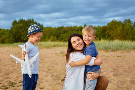 A young happy mother with two boys playing on the sand near the forest, in the hands of one of them a model of a civil aircraft and a hat on his headの写真素材