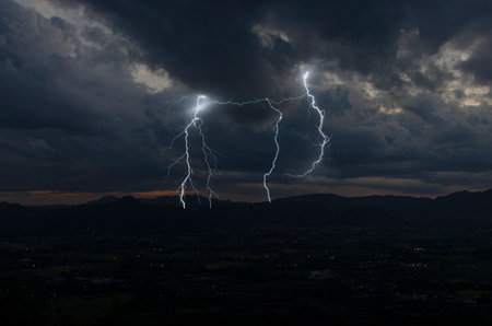 Dangerous strong lightning in black clouds over the town in the valley in the eveningの写真素材