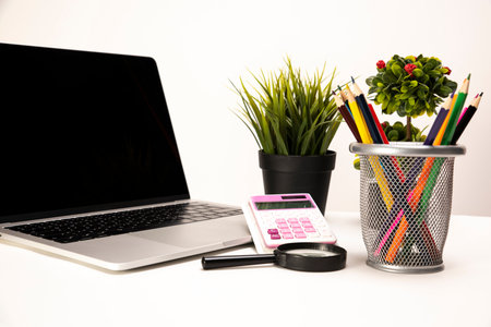 Laptop, calculator, magnifier, organizer, crayons and two green plants neatly placed on a clean white deskの写真素材