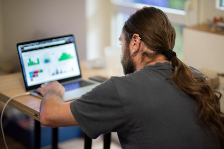 Young man with long hair is reviewing some charts on his computers screen while working in a cozy...