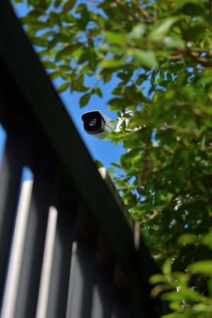 A modern white security CCTV camera on a house gate wall, surrounded by trees, ready to monitor...