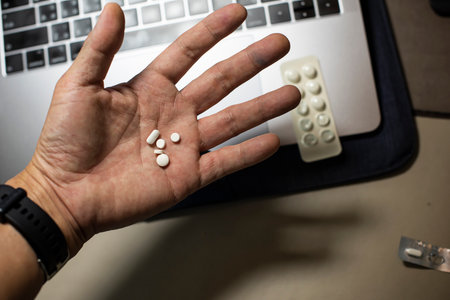 Sick male home office worker taking pills in front of his working space, during his working deadline night. Business or study with stress, late or overwork concepts. Young entrepreneur or student.の写真素材