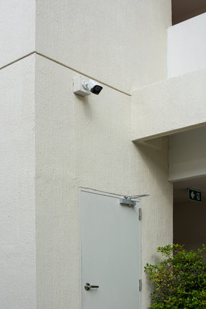 CCTV camera over the fire exit door with a white concrete wall. Security and safety system for a...