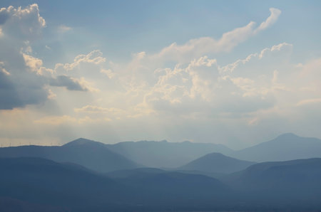 Scenic landscape of mountain ridges and sun rays getting through the cloudy skyの写真素材