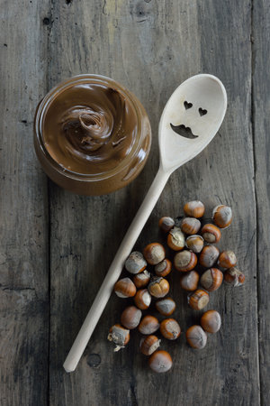 Vase with hazelnuts paste, wooden spoon with face and dry hazelnuts on wooden background
