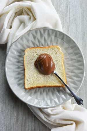 Plate with spoonful of hazelnut praline paste on slice of bread,white towel and grey table as...