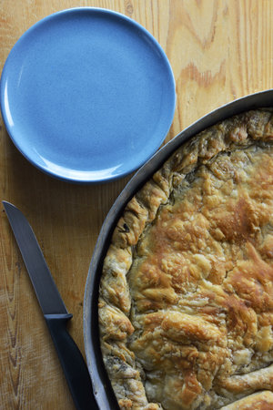 Traditional greek spinach cheese pie in baking pan, plates and knife on wooden dough rolling board