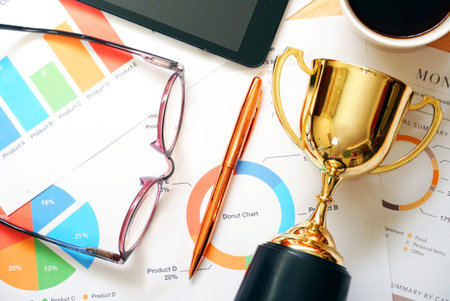 Trophy and tablet and coffee cup and pen and glasses and chart paper on working table.