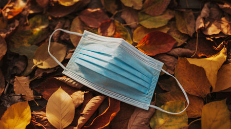 A blue disposable face mask rests on a bed of fallen autumn leaves, symbolizing the end of an era and environmental concerns.の素材