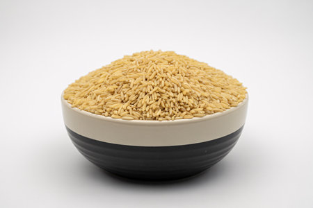 Close up shot of a black and white ceramic bowl overflowing with raw healthy brown rice grains The food is presented simply on a plain white studio backgroundの素材