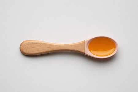 Top view of a small light brown wooden spoon holding a pool of shiny golden honey. The spoon is placed horizontally on a clean, plain white surface.の素材