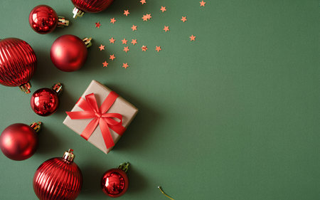 A top-down view of red Christmas ornaments and a small gift box with a red ribbon, scattered on a plain green surface with confetti.の素材