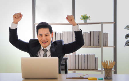 Portrait of a cheerful Asian businessman sitting at a desk looking at the PC screen, fists make him...