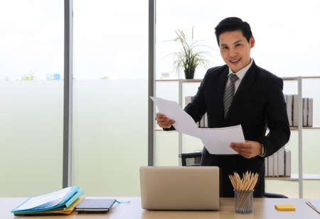 Asian businessman That is positive adults Stand up and hold documents Look at the camera and smile and relax in a modern office. Concepts that work successfully There is a computer on the desk.の写真素材