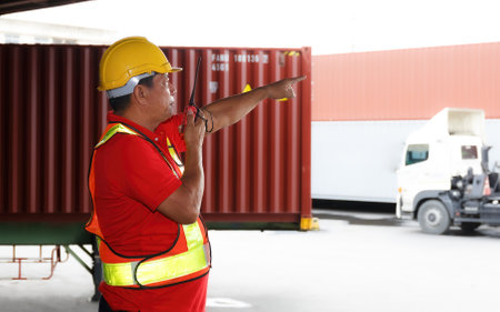 Asian male worker in safety helmet wearing hard hat Walkie Talkies Command Inventory Check with Container Background Check containers at the warehouse, import, export, logistics industry services.の写真素材