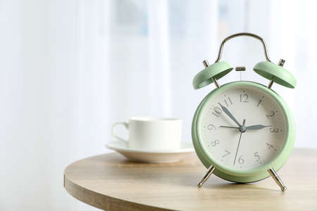 Beautiful retro alarm clock with cup of coffee on table against light background