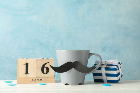 Cup with decorative mustache, gift box and wooden calendar on white table against blue background, space for textの写真素材
