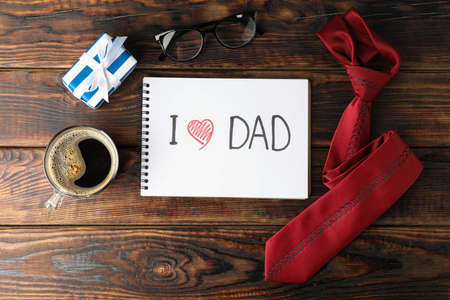 Notebook with inscription I love DAD, cup of coffee, glasses, gift box and tie on wooden background, space for text and top viewの写真素材