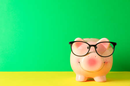 Happy piggy bank with glasses on color table against green background, space for text. Finance, saving moneyの写真素材