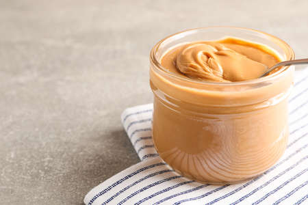 Glass jar with peanut butter and spoon, and kitchen towel on grey background, space for text and...