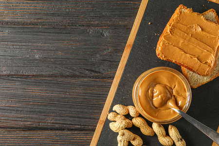Glass jar with peanut butter and spoon, peanut, peanut butter sandwich and cutting board on wooden...