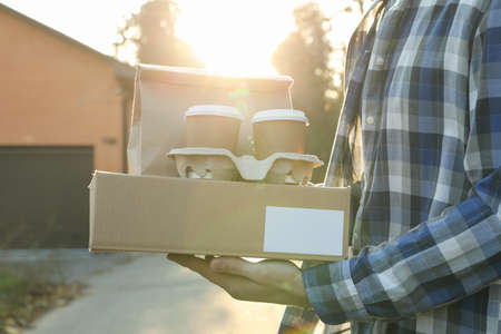 Man holds blank box, coffee cups and paper package outdoor. Delivery