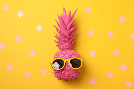 Painted pink pineapple with sunglasses on decorated yellow background, top view
