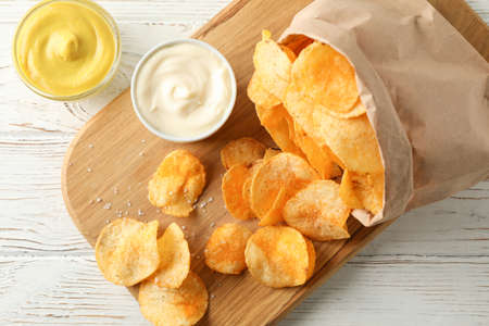 Paper bag of potato chips. Beer snacks, sauce on cutting board, on white wooden background, space...