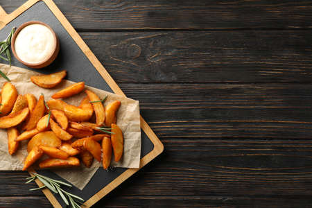 Slices of baked potato wedges, rosemary, white sauce, kitchen board on wooden background, space for...