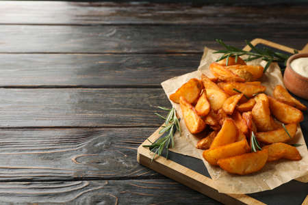 Slices of baked potato wedges, rosemary, white sauce, kitchen board on wooden background, space for...