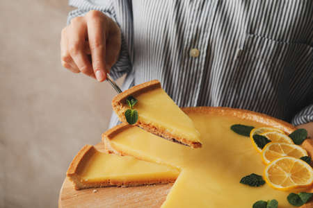 Woman holding wooden board and spatula with lemon tart against brown background, close up