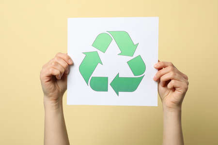 Female hands holding sheet with recycle sign against beige background, close up